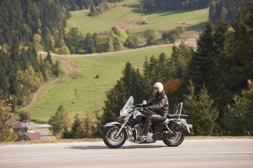 Fototapeta na wymiar Side view of bearded motorcyclist in helmet, sunglasses and black leather clothing riding cruiser motorbike along narrow asphalt path on sunny summer day on background of tall trees and greeen grass.