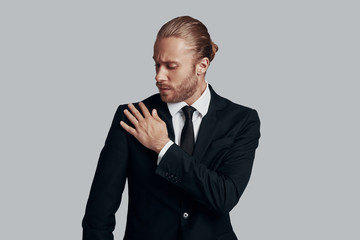 Checking details. Handsome young man in full suit cleaning jacket while standing against grey background