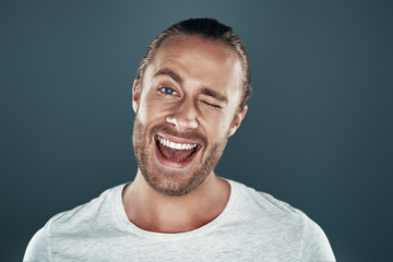 Winking. Handsome young man looking at camera and smiling while standing against grey background