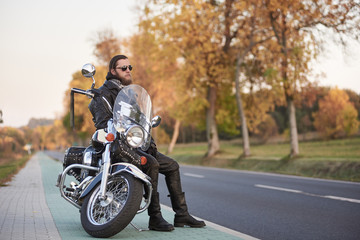 Young bearded biker in black leather clothing and dark sunglasses sitting on motorcycle on clean paved roadside, on background of empty straight asphalt road and vintage trees golden bokeh foliage.