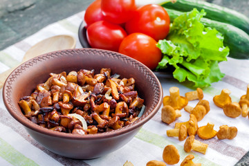 Fried chanterelles with onion in rustic bowl and plate with fresh vegetables for salad on background
