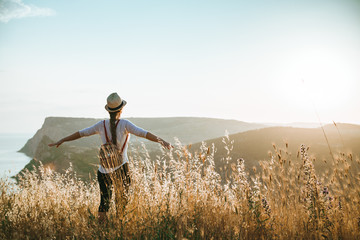 A young girl stands with a hat and a backpack against the backdrop of mountains bathed in the rays of the sun. Summer background with place for your text