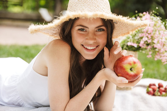 Photo of beautiful young woman wearing straw hat eating apple while having picnic in green park