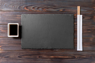 Empty rectangular black slate plate with chopsticks for sushi and soy sauce on wooden background. Top view with copy space