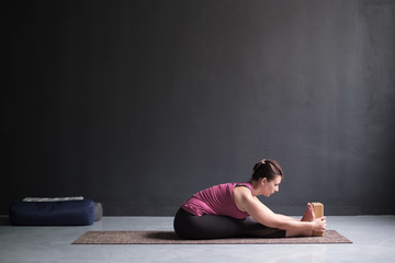Woman practicing yoga, doing paschimottanasana exercise, Seated forward bend pose, working out. Indoor full length, black yoga studio