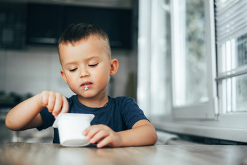 Cute boy in the kitchen eating yogurt from a white container for yogurt, there is a place for advertising