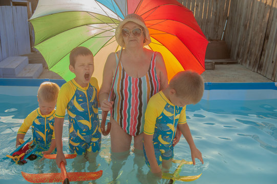 Elderly woman in striped swimsuit, sunglasses and elegant hat with rainbow color umbrella standing in pool. Children in swimwear swim next to their grandmother and play with bright plastic airplanes