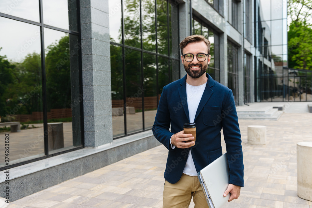 Wall mural Portrait of smiling businessman wearing eyeglasses holding laptop and paper cup