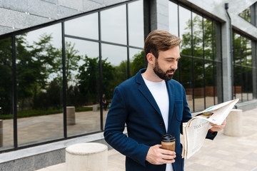 Portrait of confident businessman in jacket drinking coffee from paper cup and reading newspaper...