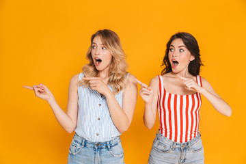 Portrait of two excited blonde and brunette women 20s in summer wear smiling while pointing fingers at copyspace