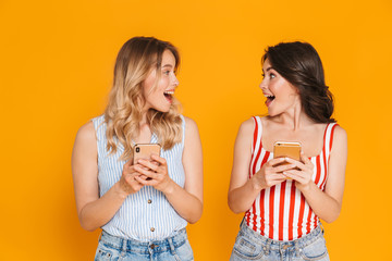 Portrait of two delighted blonde and brunette women 20s in summer wear smiling while holding cellphones