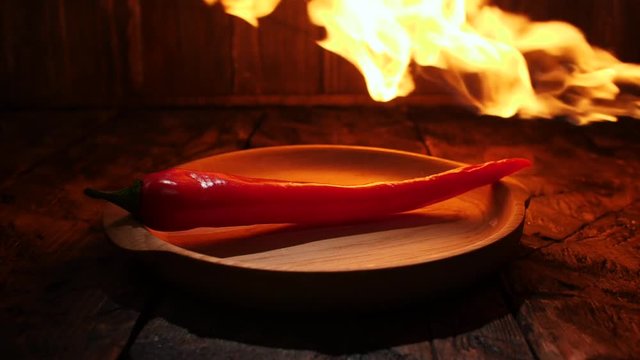 Close-up view of red hot chilli pepper laying in the wooden plate on the wooden table with jet of flame on the background. Stock footage. Spicy food concept