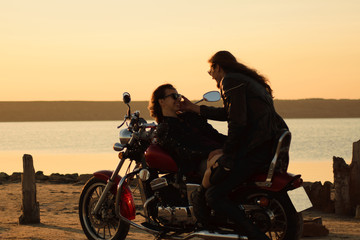 Fototapeta na wymiar Romantic biker couple in leather and sunglasses with red motorcycle. Handsome bearded long-haired man and attractive woman outdoors against motorcycle and sunset, sunrise
