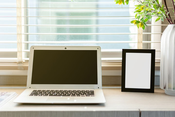 Fototapeta premium Blank black color picture frame template for place image or text inside on the desktop table work space with laptop and flower. window light