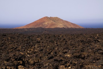 View over rough volcanic lava rocks on red cone of volcano with blurred atlantic ocean background -...