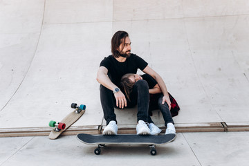 Young father and his son dressed in the casual clothes sit in an embrace at the slide in a skate park next to the skateboards at the sunny day