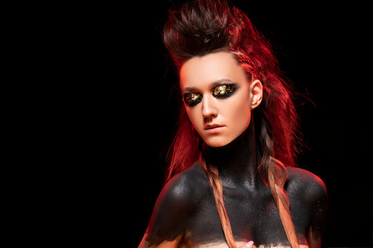 Portrait of a young savage girl. Naked shoulders and neck are covered with black paint. Conceptual makeup with gold leaf on her eyelids. Black background. Fashion and commercial design