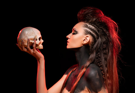 Portrait of a young savage girl who holds a skull in her hand. Naked shoulders and neck are covered with black paint. Conceptual makeup with gold leaf on her eyelids. Black background.