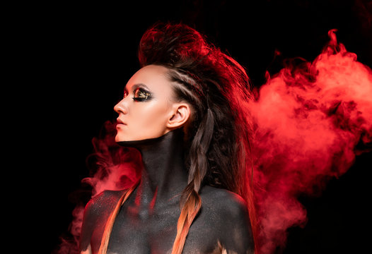 Portrait of a young savage girl in red smoke. Naked shoulders and neck are covered with black paint. Conceptual makeup with gold leaf on her eyelids. Black background. Commercial design.