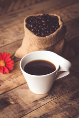 Black coffee In the cup white have roasted coffee beans are In a cloth bag sack and on table with, still have red flowers and stainless steel spoon all placed on a wooden table.