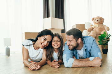Fototapeta na wymiar Happy family having fun while moving into new apartment. Mother, father and daughter together smiling, with Cardboard boxes on background