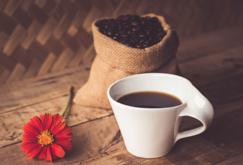 Black coffee In the cup white have roasted coffee beans are In a cloth bag sack and on table with, still have red flowers and stainless steel spoon all placed on a wooden table.