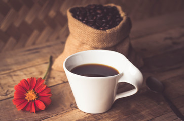 Black coffee In the cup white have roasted coffee beans are In a cloth bag sack and on table with, still have red flowers and stainless steel spoon all placed on a wooden table.