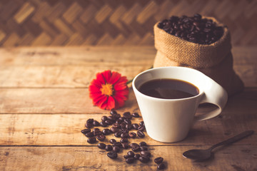 Black coffee In the cup white have roasted coffee beans are In a cloth bag sack and on table with, still have red flowers and stainless steel spoon all placed on a wooden table.