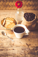 Black coffee In the cup white have roasted coffee beans are In a cloth bag sack butter cracker In the basket weave bamboo, still have red flowers and stainless steel spoon all placed on a wooden table