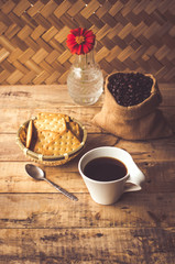 Black coffee In the cup white have roasted coffee beans are In a cloth bag sack butter cracker In the basket weave bamboo, still have red flowers and stainless steel spoon all placed on a wooden table