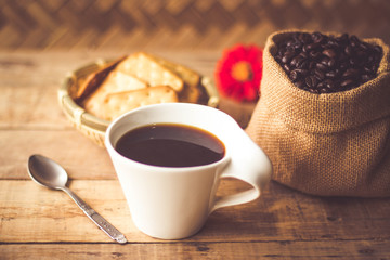 Black coffee In the cup white have roasted coffee beans are In a cloth bag sack butter cracker In the basket weave bamboo, still have red flowers and stainless steel spoon all placed on a wooden table