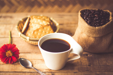 Black coffee In the cup white have roasted coffee beans are In a cloth bag sack butter cracker In the basket weave bamboo, still have red flowers and stainless steel spoon all placed on a wooden table