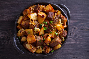 Fried beef and  potatoes with onions and garlic served in black dish on wooden background