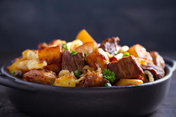 Fried beef and  potatoes with onions and garlic served in black dish on wooden background