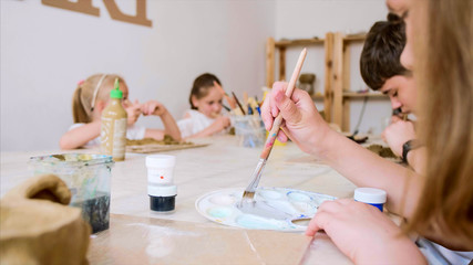 Children are sitting at table in workshop at art lesson and modeling workpiece from wet clay.