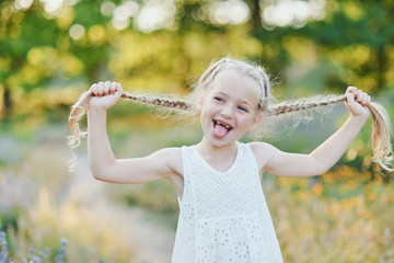 Funny girl holding hair plaits. Small cute girl with long blonde hair showing tongue outdoor. Kid...