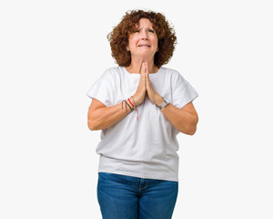Beautiful middle ager senior woman wearing white t-shirt over isolated background begging and...