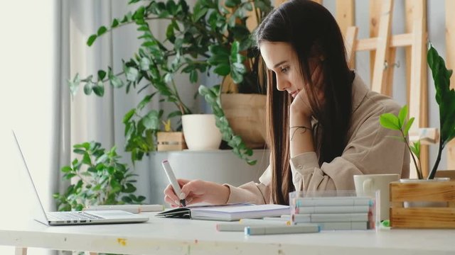 Side view of a beautiful young woman illustrator making a new work for an illustration of a children's book sitting at a table with a laptop. The concept of creating a unique art