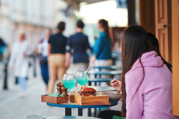 Young woman eating delicious grilled burger and enjoying while sitting outdoor. Girlfriends relaxing on street cafe terrace with alcoholic cocktails and black burgers in wooden food presentation crate