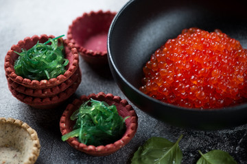 Closeup of a black bowl with red caviar and vegetable tartlets with chuka salad, selective focus