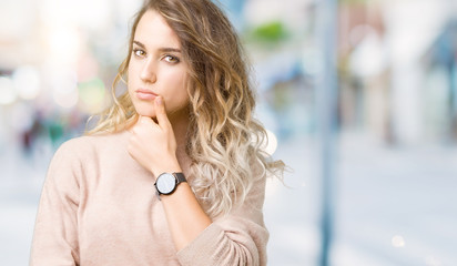 Beautiful young blonde woman wearing sweatershirt over isolated background looking confident at the camera with smile with crossed arms and hand raised on chin. Thinking positive.