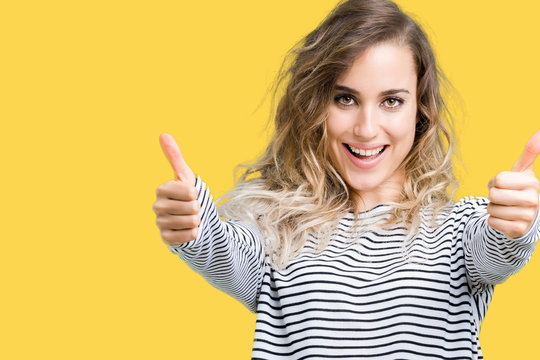 Beautiful young blonde woman wearing stripes sweater over isolated background approving doing positive gesture with hand, thumbs up smiling and happy for success. Looking at the camera