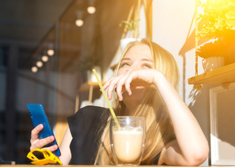 happy smiling young blond woman drinking a latte in a cafe for a glass, sunglasses and phone in the foreground. morning sun, bright summer day