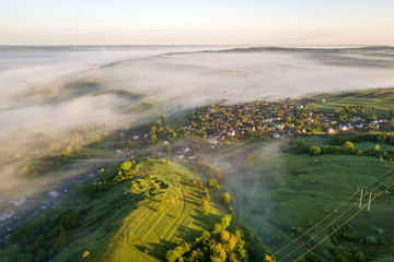 Top view of foggy green grassy hill, village house roofs in valley among green trees on blue sky background. Spring misty landscape panorama at dawn.