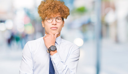 Young handsome business man with afro wearing glasses looking confident at the camera with smile with crossed arms and hand raised on chin. Thinking positive.