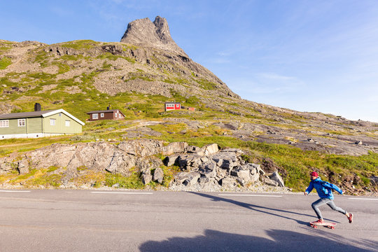 Trollstigen, Romsdalen, Norway: A man on his longboard cruising u on the narrow pass road with typical Scandinavian cabins in the background.