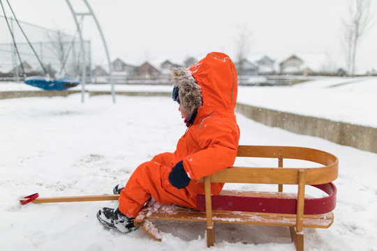 Toddler boy sitting on wooden sled at snow covered park.