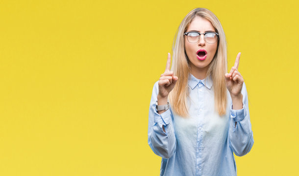 Young beautiful blonde business woman wearing glasses over isolated background amazed and surprised looking up and pointing with fingers and raised arms.