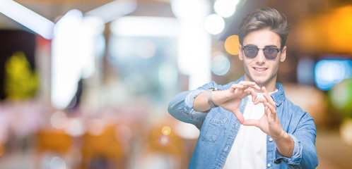 Young handsome man wearing sunglasses over isolated background smiling in love showing heart symbol and shape with hands. Romantic concept.