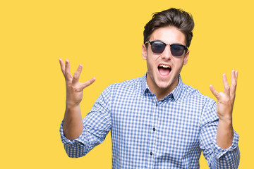 Naklejka premium Young handsome man wearing sunglasses over isolated background crazy and mad shouting and yelling with aggressive expression and arms raised. Frustration concept.